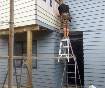 Worker standing on the top step of a stepladder balanced on a scaffold platform to reach cladding an unsafe working-at-height practice