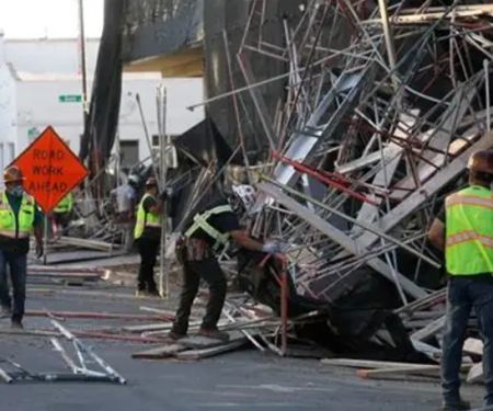Workers in hi-vis vests clearing debris from a scaffold collapse across a town street, with a "Road Work Ahead" sign in the foreground